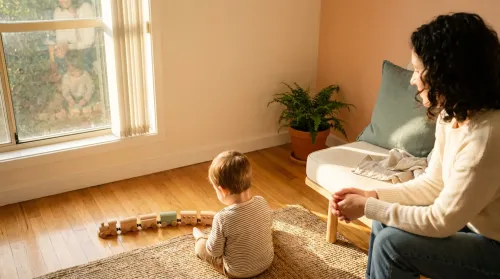 Toddler arranges wooden trains in a row on a jute rug while a parent watches from a cream couch in a sunlit room